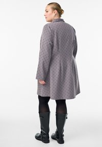 Nainen, jolla on vaaleat hiukset nutturalla, pukeutuneena harmaaseen pilkulliseen takkiin, mustiin sukkahousuihin ja mustiin polvipitkiin saappaisiin, seisoo kyljellään valkoista taustaa vasten.