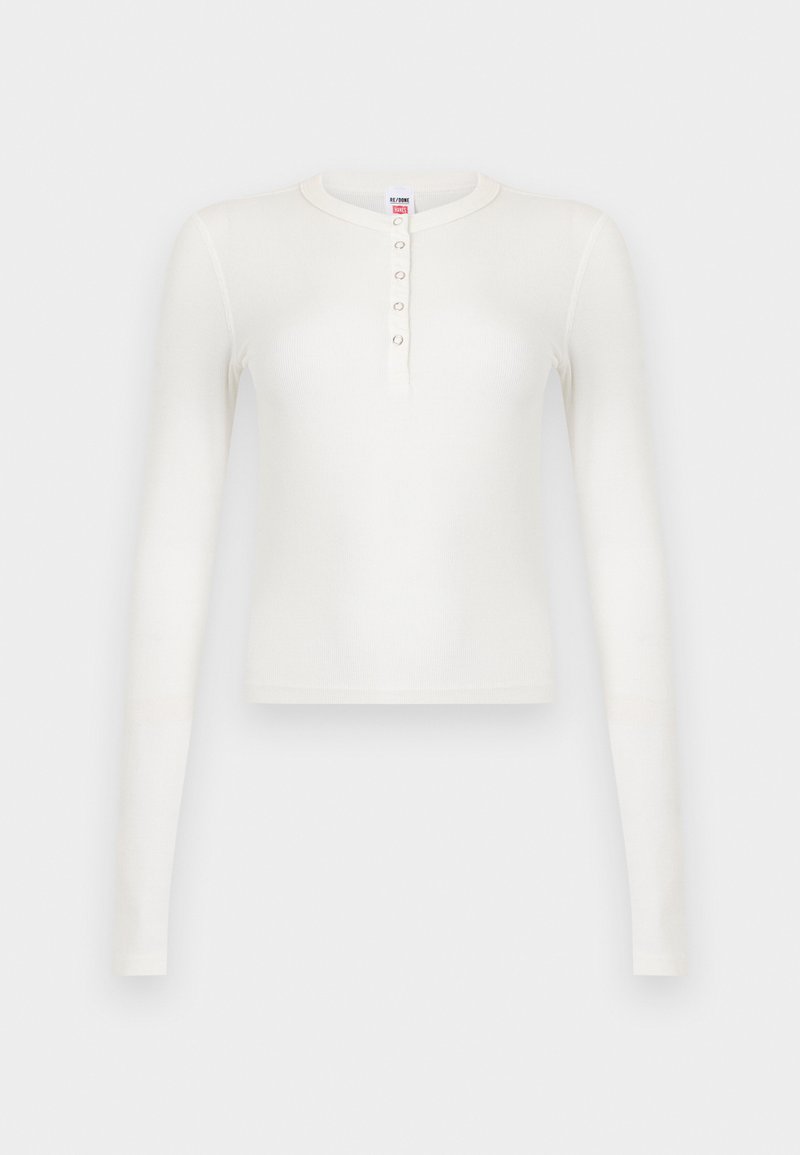 Haut blanc à manches longues en tricot côtelé avec col rond et patte de boutonnage munie de cinq petits boutons, présenté sur un fond blanc.