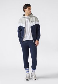 Uomo che indossa pantaloni joggers blu navy, t-shirt bianca, felpa con zip grigia e blu navy, calze bianche e blu a righe, e scarpe da ginnastica bianche, in piedi su uno sfondo bianco.
