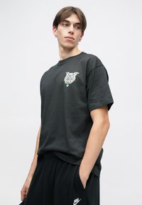 Μαύρη βαμβακερή φανέλα με κοντά μανίκια, με γραφικό σχέδιο στο αριστερό στήθος. Φοριέται με μαύρη βερμούδα και σε χαλαρή γραμμή.