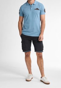 Vaaleansininen polo-paita lyhyillä hihoilla, jossa on rintatasku, yhdistetty mustiin cargo-shorteihin ja valkoisiin tennareihin.