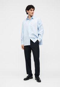 Mężczyzna w jasnoniebieskiej, luźnej koszuli, czarnych spodniach i czarnych eleganckich butach, stoi z jedną ręką w kieszeni, patrząc na bok.