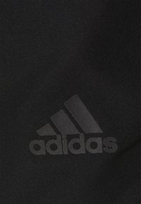 Černá tkanina s hladkou texturou, na které je výrazně a centrálně umístěno vystouplé šedé logo Adidas a tři diagonální pruhy.