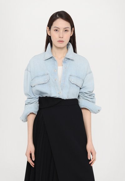Camicia in denim azzurro chiaro con colletto e tasche frontali, abbinata a una gonna a pieghe nera avvolgente. La camicia ha le maniche arrotolate.