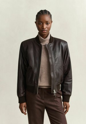 Femme de face portant une veste en cuir marron foncé, un col roulé beige et un pantalon marron, sur un fond uni et clair.