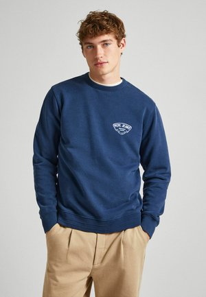 Tamsiai mėlynas džemperis su raibu apykakle ir juosmeniu, turintis baltą logotipą ant krūtinės. Minkšta medvilnė ir laisvas kirpimas.