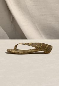 Καφέ φλατ σανδάλι με τύπωμα φιδιού, χαμηλό τακούνι και λουράκι ανάμεσα στα δάχτυλα, τοποθετημένο σε ανοιχτό μπεζ επιφάνεια με υφασμάτινο φόντο.