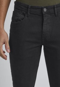 Jeans de mezclilla negra con un corte ajustado. Las características incluyen un cierre delantero con botón, presillas para cinturón y bolsillos frontales. Textura suave y mate.