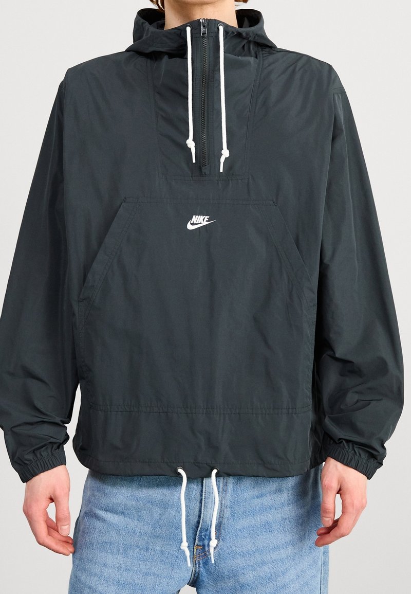 Personne portant une veste coupe-vent noire à demi-zip de Nike avec des cordons blancs et une poche avant, associée à un jean bleu clair.