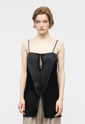 Mladá žena s krátkymi vlasmi nosí čierny saténový top bez rukávov s tenkými ramienkami a detailom zaväzovania vpredu, v kombinácii s béžovými nohavicami.