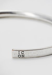 Sølvmansjettarmbånd med en glatt, avrundet design, med graverte bokstaver "IC ON" i den ene enden. Polert overflate med en lett tekstur.