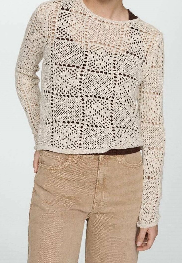 Beige gehaakte trui met lange mouwen en geometrische vierkantpatronen, gedragen over een donker shirt, gecombineerd met lichtbruine, broek met hoge taille.