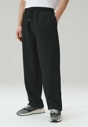 Person trägt schwarze Jogginghose mit Kordelzug und graue Turnschuhe, steht mit den Händen in den Taschen vor schlichtem Hintergrund.