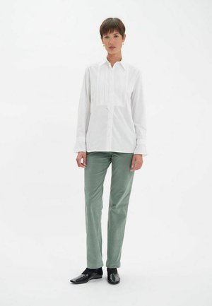 Chemise blanche à manches longues avec des plis verticaux et fermeture à boutons, associée à un pantalon vert clair et des chaussures noires, se tenant devant un fond blanc.