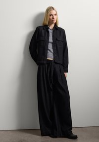 Giacca in denim nera e pantaloni a gamba larga abbinati a un top corto in maglia grigia, caratterizzato da due tasche sul petto e un fit rilassato.