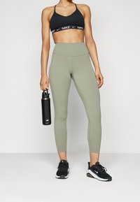 Czarny sportowy stanik z logo, wysokie oliwkowe legginsy, czarna butelka na wodę i czarne sneakersy z białymi akcentami, stojące na tle w neutralnych kolorach.