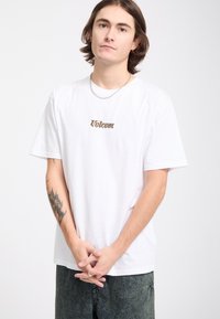 Jeune homme aux cheveux bruns mi-longs porte un t-shirt blanc Volcom et un jean foncé, les mains jointes devant, sur un fond clair uni.