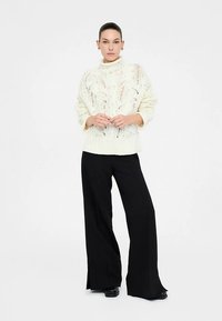 Kremowy sweter z wysokim kołnierzem i ażurowym wzorem, zestawiony z czarnymi szerokimi spodniami, tworząc kontrast warstwowej tekstury.