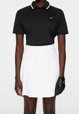 Zwart poloshirt met witte strepen op de kraag, gecombineerd met een witte geplooide rok. Zwarte handschoen met logodetail aan de rechterhand.