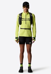 Camiseta amarilla brillante de manga larga con tejido texturizado, pantalones cortos negros y chaleco de hidratación. Gorra negra y gafas de sol; acentos verdes vibrantes en los guantes y calcetines.