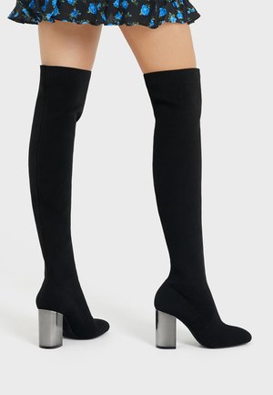 Femme portant des bottes noires montantes avec des talons blocs argentés et une jupe courte noire avec un motif floral bleu sur fond blanc.