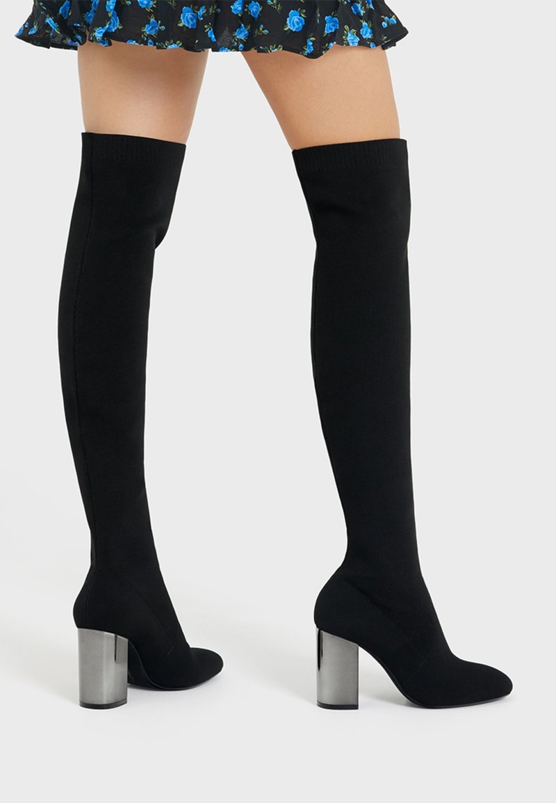 Femme portant des bottes noires montantes avec des talons blocs argentés et une jupe courte noire avec un motif floral bleu sur fond blanc.