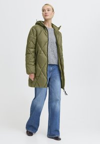 Olijfgroene gevoerde jas met capuchon, met een gewatteerd ontwerp en zijzakken, gedragen over een grijze trui en blauwe wijde jeans.