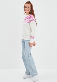 Witte gebreide trui met een roze geometrisch patroon bij de halslijn, gecombineerd met lichtblauwe jeans en witte platform sneakers.