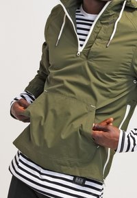 Osoba w oliwkowej kurtce z pół-zamkiem nosi czarną i białą koszulę w paski, trzymając ręce otwarte przednią kieszeń kangurkę.