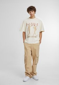 Kremo spalvos grafiškas t-shirt su per dideliu fasonu, papuoštas dviejų figūrų atspaudu ir ryškiu užrašu; derinamas su šviesiai smėliniais kargo džinsais ir baltais sportiniais bateliais.