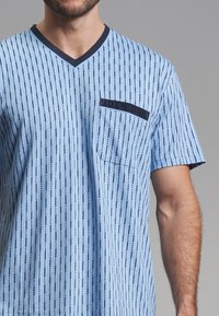 Camisa de manga corta azul claro con rayas verticales, cuello en V y un bolsillo en el pecho izquierdo con un acabado oscuro en contraste. Textura de tela suave.