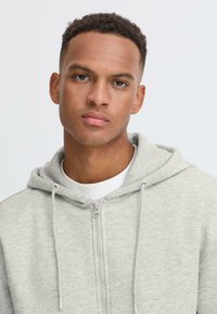 Gray zip-up hoodie with drawstrings, featuring a soft texture. Layered over a white crew-neck shirt, against a neutral background.