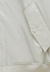 Tissu beige clair avec une poche à rabat cousue sécurisée par un bouton-pression, et une ceinture élastique côtelée en bas.