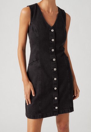 Robe en denim noire sans manches avec boutons métalliques sur le devant, encolure en V, poches latérales, portée par une personne debout avec les bras détendus.