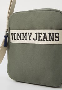 Borsa a tracolla verde in tessuto con una banda crema prominente con "TOMMY JEANS" in lettere nere. Inclusa tracolla regolabile.