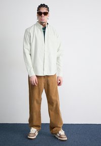 Lys grøn langærmet skjorte, brune fløjl-bukser og beige sneakers. Tilbehør inkluderer mørke solbriller og en kædehalskæde.