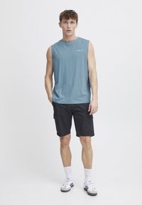 Canotta azzurro chiaro senza maniche con la scritta "A.R.C." sul petto, abbinata a pantaloni cargo neri. Il modello indossa calzini bianchi e sneakers nere e grigie.
