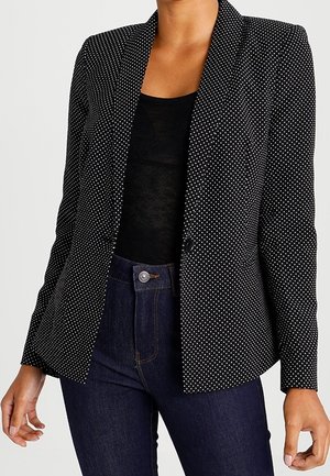 Vrouw die een zwart polka-dot blazer draagt over een zwarte top, gecombineerd met een high-waisted donkerblauwe jeans, staand tegen een effen achtergrond.