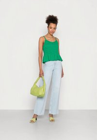 Top verde com plissado e peplum, com alças finas, combinado com jeans de perna larga azul claro, mala verde e saltos a condizer, apresentado contra um fundo neutro.