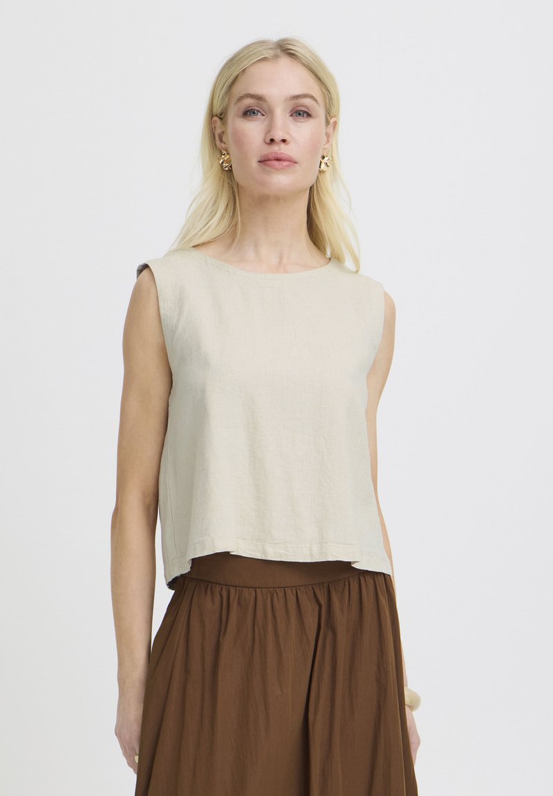 Femme blonde portant un haut beige sans manches, une jupe marron taille haute et des boucles d'oreilles dorées, debout devant un fond clair uni.