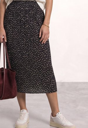 Femme portant une jupe plissée noire à pois blancs, des baskets blanches, un bracelet en or et tenant un sac cabas en cuir rouge foncé.