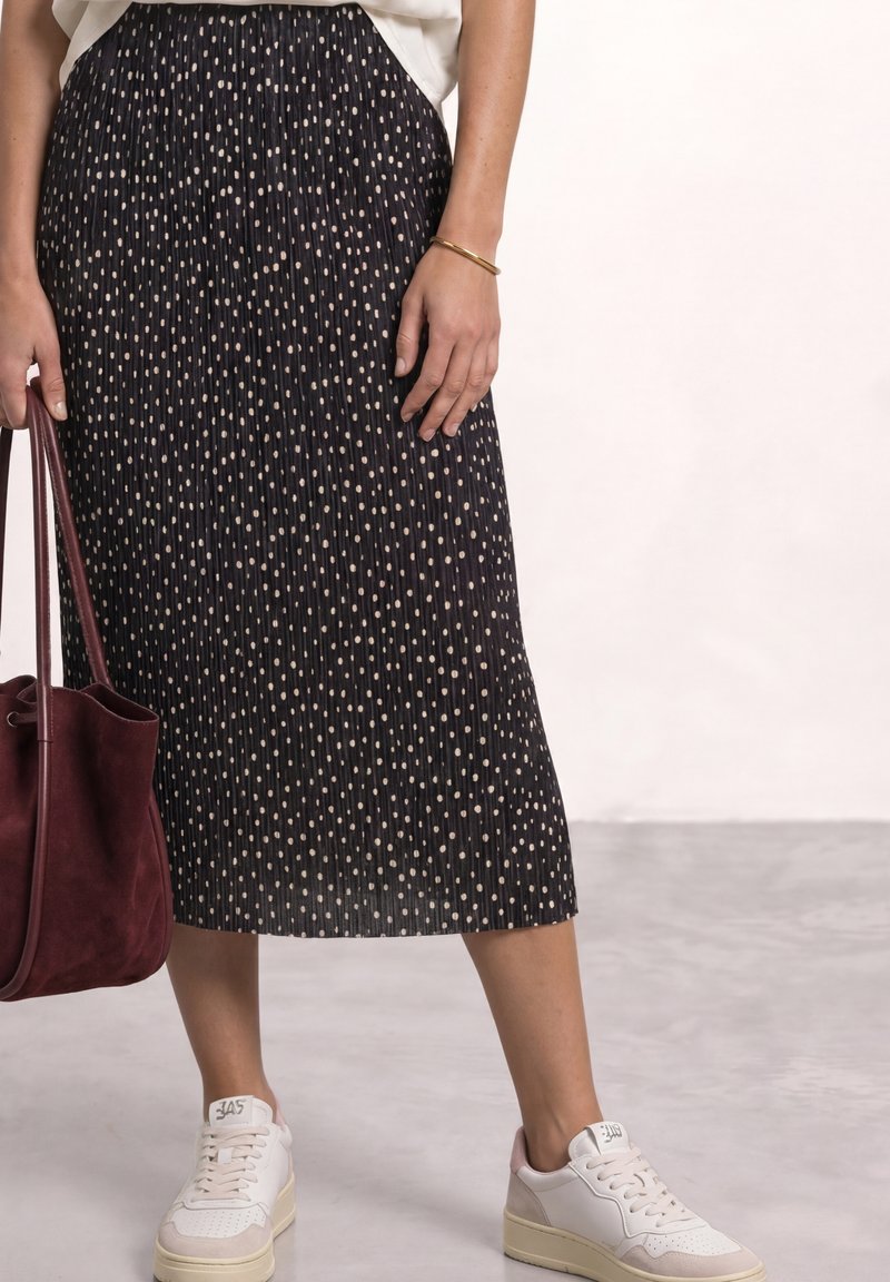 Woman wearing a black pleated skirt with white polka dots, white sneakers, a gold bracelet, and holding a dark red leather tote bag.