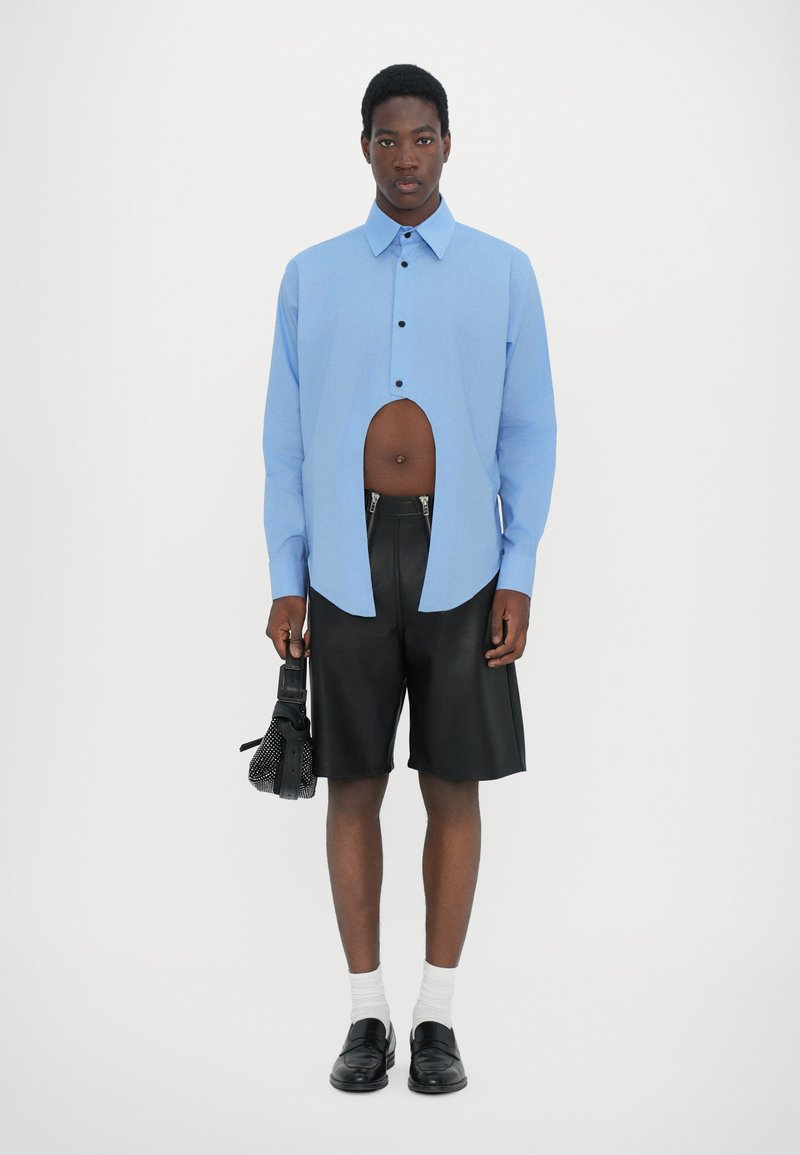 Lysblå knapp-up skjorte med udskæringsdetalje, parret med sorte lædershorts, hvide sokker og sorte sko, mens der holdes en lille taske.