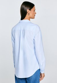 Sieviete ar taisniem tumšiem matiem valkā gaiši zilu pogājamu kreklu un zilas džinsas, skatās sāniski ar pilnībā redzamu muguru.