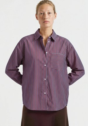 Jeune personne aux cheveux clairs porte une chemise ample rayée bordeaux et bleue avec une poche poitrine, sur un fond uni.