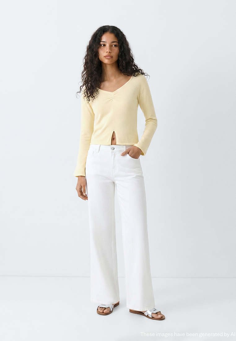 Femme aux cheveux bouclés portant un haut à manches longues jaune clair et un pantalon blanc à jambes larges avec des sandales blanches, debout devant un fond uni.