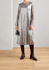 Robe en sequins argentés avec des manches longues, coupe ample, longueur genoux. Associée à des bottes en cuir noir jusqu'aux genoux, texture lisse, bout carré.