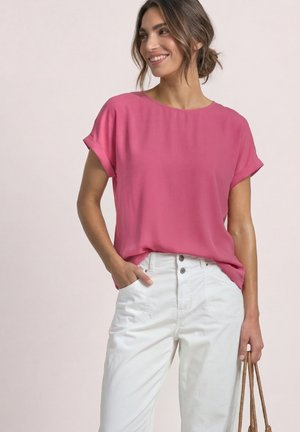 Donna sorridente che indossa una camicetta rosa a maniche corte e pantaloni bianchi a vita alta, che tiene in una mano una borsa con manici in corda.