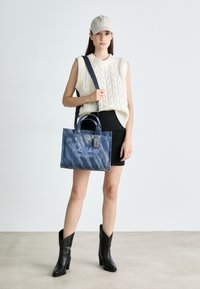Denim torba za prosti čas z modrim gradientom in vezenim napisom "Coach New York". Ima dvojne ročaje, odstranljiv trak in oznako.
