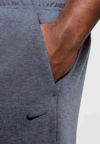 Graue Sporthose mit glatter Textur, ausgestattet mit einer Seitentasche und einem kleinen schwarzen Nike-Logo an der Vorderseite in der Nähe der Taschenöffnung.